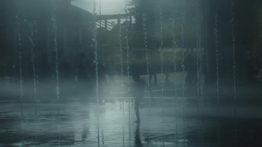A dark silhouette of a person in shorts walks slowly through the splashing water of a fountain on a damp foggy day in an urban park