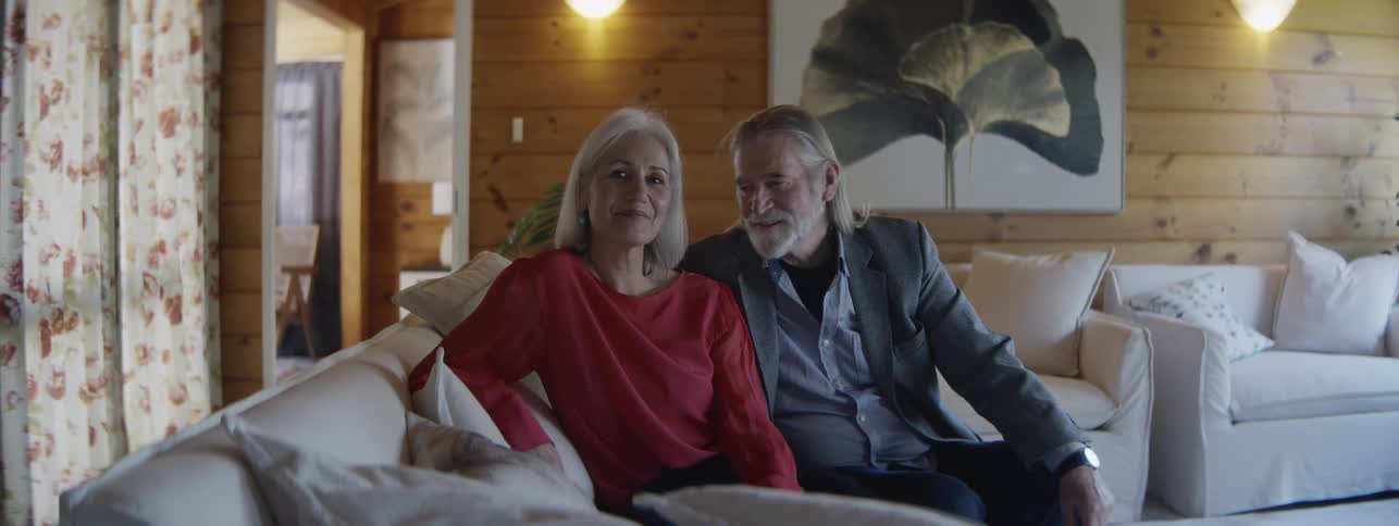 An older man with a long white beard wearing a grey blazer and an older woman with white hair wearing a coral shirt smile while sitting together on a white couch in a woodpaneled living room
