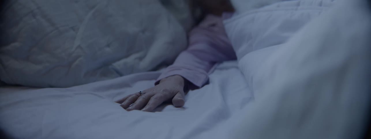 A close view shows an elderly persons hand wearing a ring resting on a white sheet before slowly withdrawing under the blankets in a dimly lit dark bluetoned bedroom