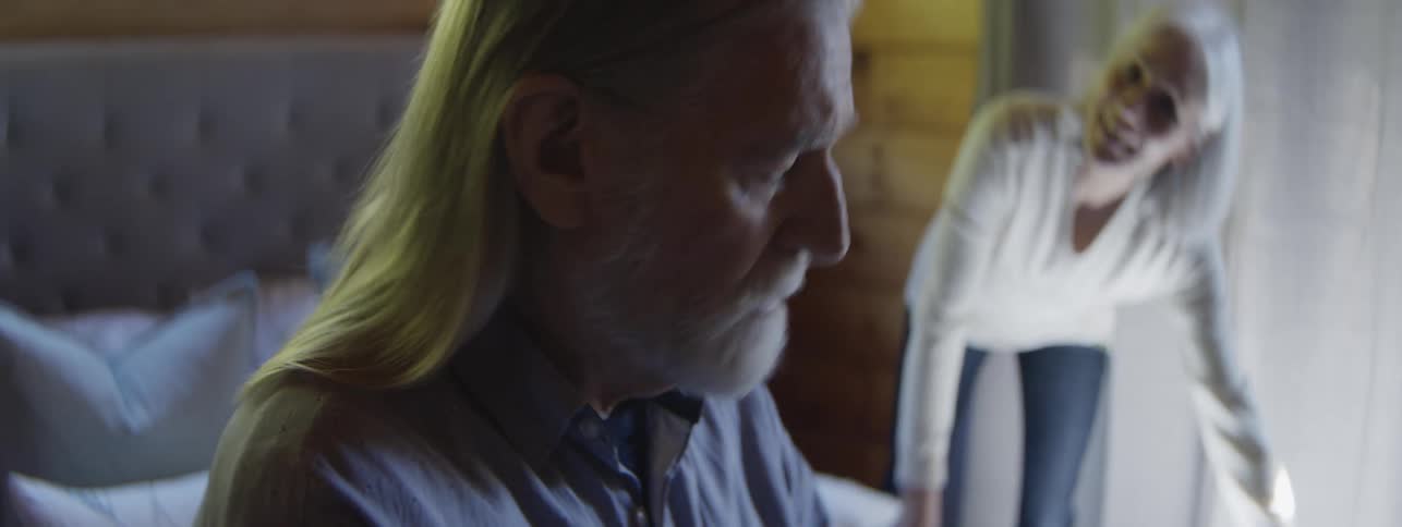 The profile of an older man with long gray hair and a beard while a woman with gray hair stands out of focus in the background and leans over a surface and they start arguing