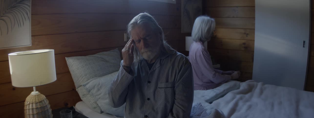 Man sitting on the edge of the bed with his hand on his forehead appearing unwell while a woman is in the background