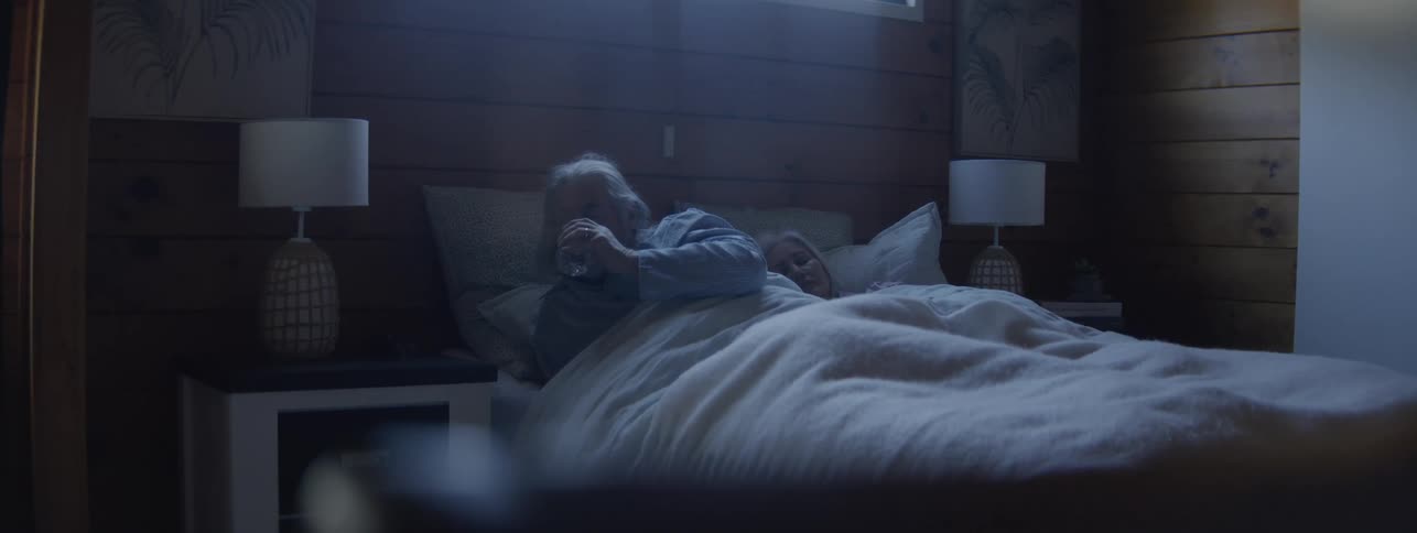 The video is a wide lowlight shot of an elderly couple asleep in a large bed in a woodpaneled roomthe woman wakes up turns over and reaches to gently touch the arm of her sleeping partner before settling back down to sleep