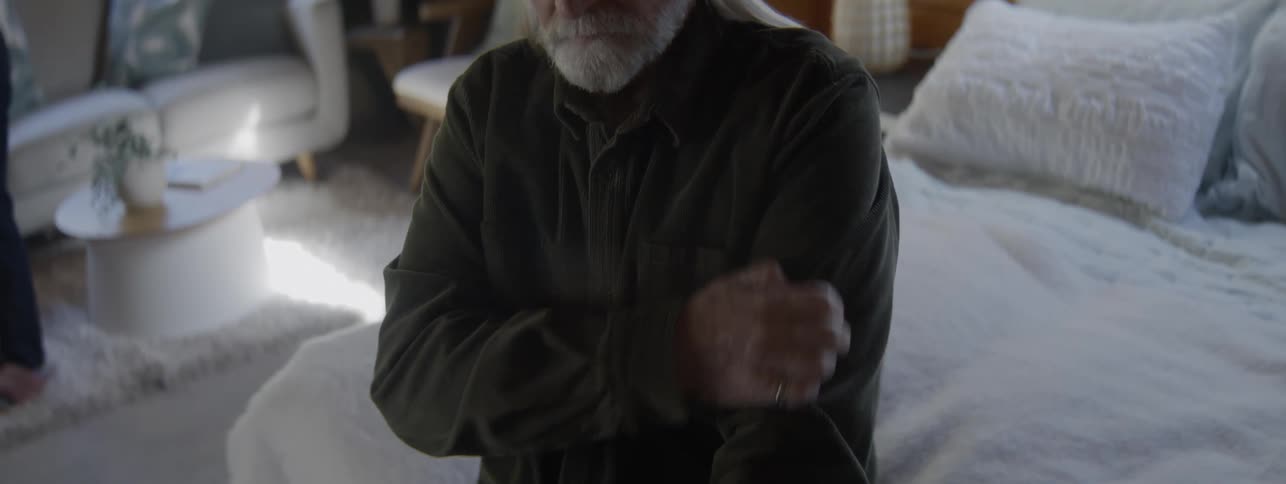 A senior man with long gray hair and a dark shirt sits in a bright modern bedroom with white furniture and rubs his shoulder and chest area as if in discomfort