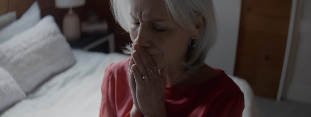 A close view shows an older woman with white hair wearing a coral shirt who brings her hands to her face in distress while sitting on a bed in a dimly lit bedroom