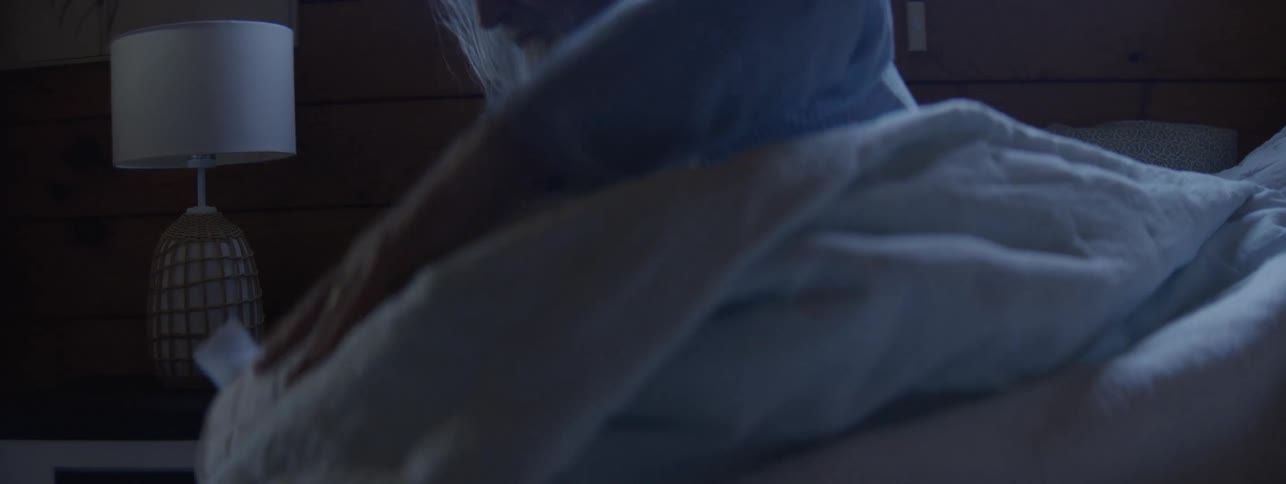The video is a lowlight interior shot of a man sitting up in bed next to a woman who is still sleepinghe smooths the duvet and adjusts the bed covers on his side before settling back down under the blankets