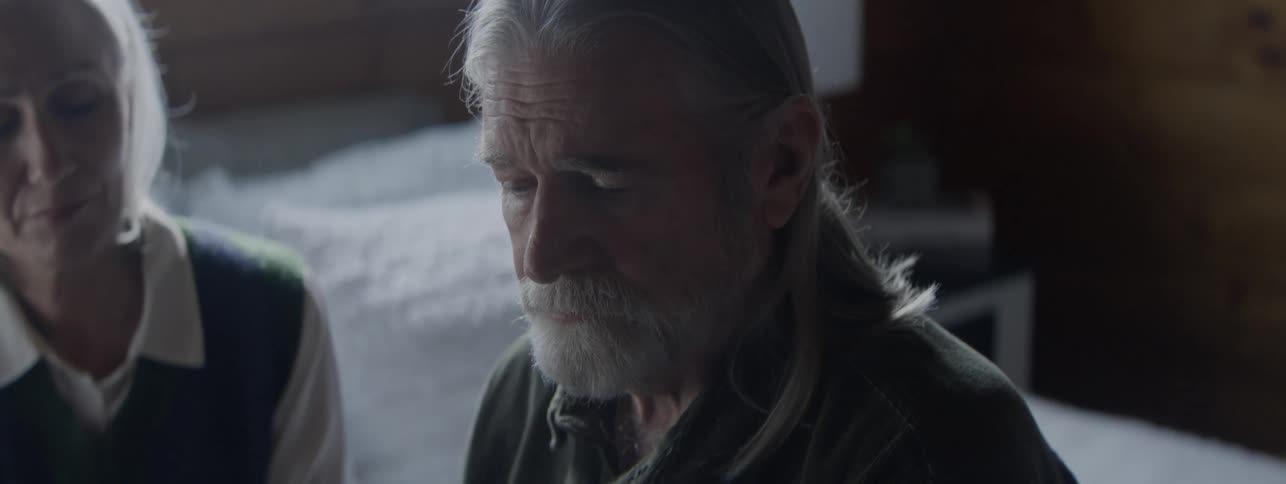 The video is a closeup of an older man with a long gray beard and shoulderlength gray hairhe is seated and looking down with a somber or thoughtful expression in a lowlight indoor setting