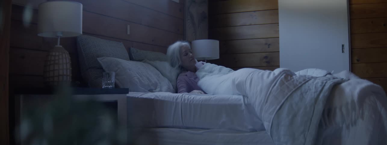 An older woman with white hair in pink pajamas wakes up abruptly in the morning sitting up from a white bed in a dark woodpaneled bedroom