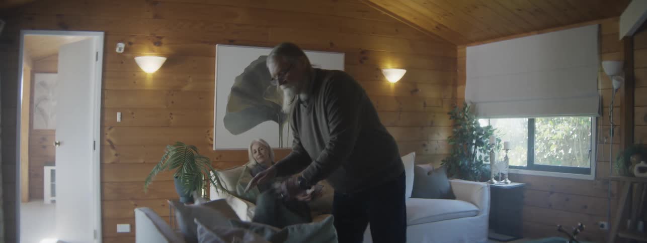 An older man with long hair and a beard walking around a woodpaneled living room picking up an object from a table straightening pillows on the couch where an older woman is reading and then turning as the woman stands up