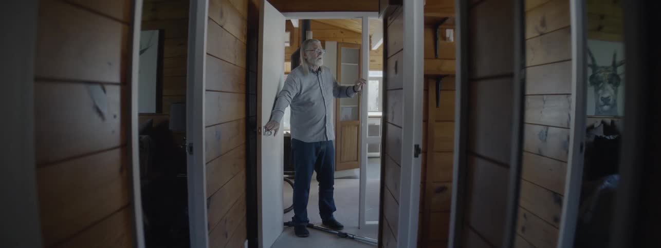 An older man with long grey hair and glasses struggles to vacuum a carpeted hallway stopping the chore to cough and catch his breath in a woodpaneled interior