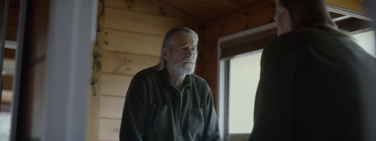 An elderly confused man with dementia looking in the mirror of a wooden house at dawn