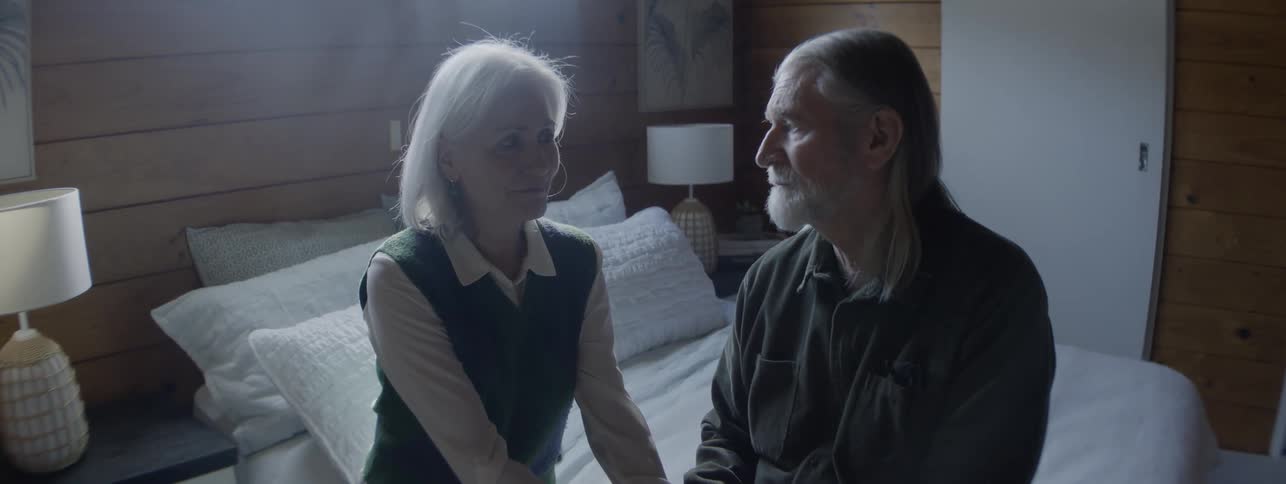 A senior man with long gray hair and a dark shirt sits on a bright bed in a woodpaneled room next to a senior woman in a green sweater vest as they talk intently and hold hands