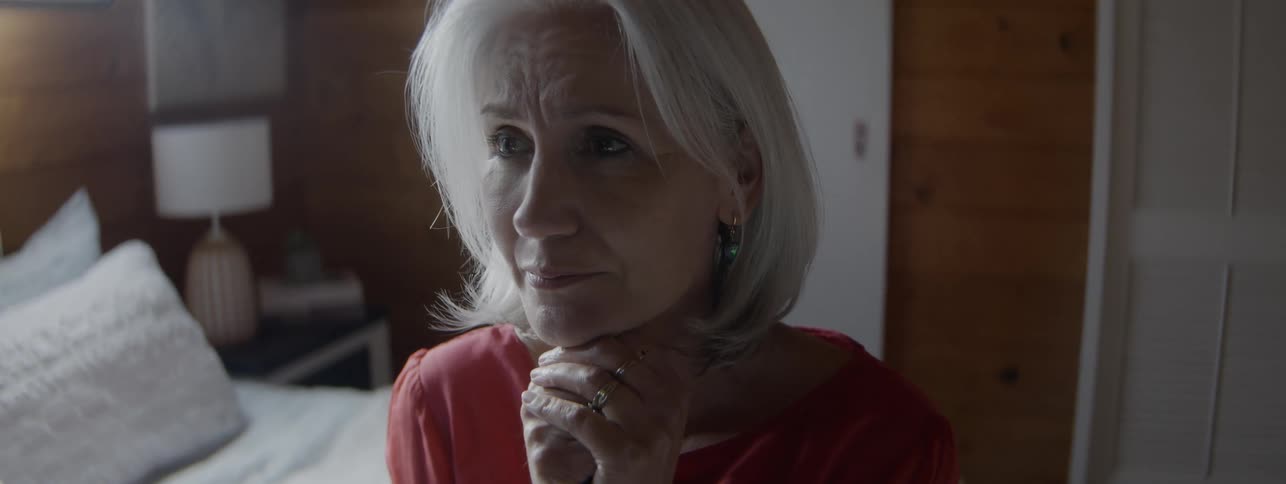 An older person sits in a room with their hands clasped near their chin
