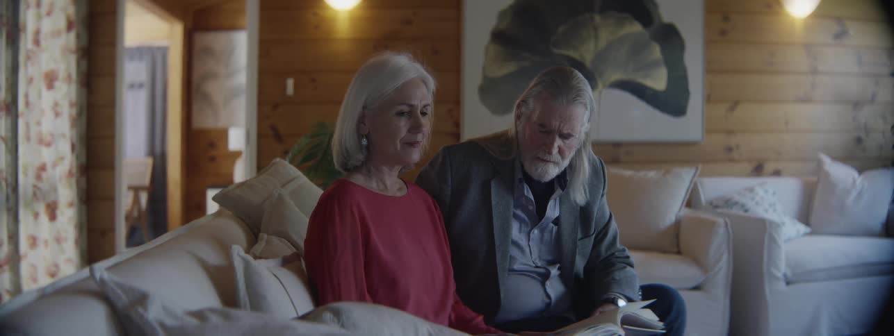 A man and a woman with gray hair sitting on a couch looking at a book that the woman is holding and pointing to and the woman is smiling