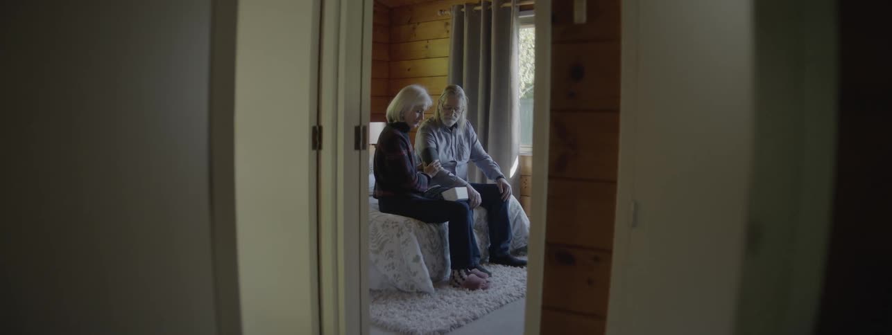 An older man and older woman with white hair are seated on a bed in a woodpaneled room as the woman wraps a blood pressure cuff around the mans arm