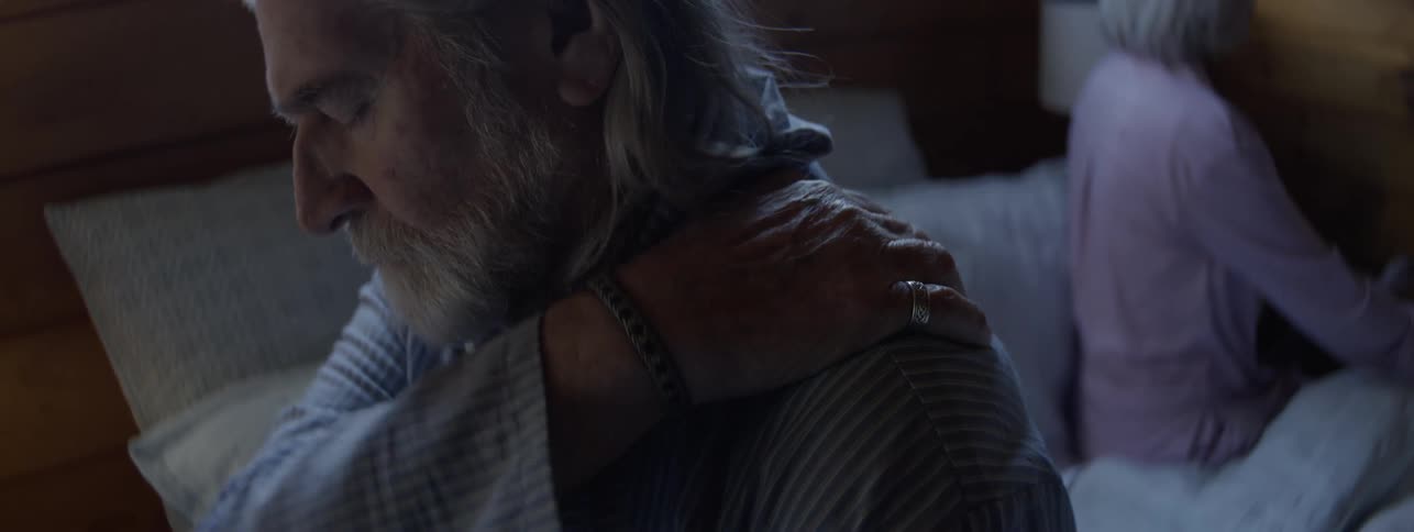 An elderly man with a long white beard and gray hair wearing a striped shirt sitting up in a dimly lit bedroom setting with a woman in the background