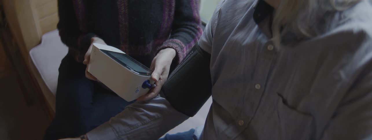 A person securing a black cuff around the bicep of another person then holding the white blood pressure monitoring device and pushing a button to begin the measuring process