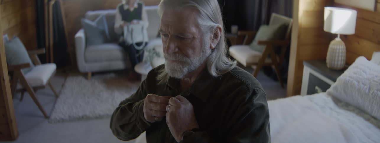 Elderly man with arthritis struggles to button up his shirt while getting ready in a room