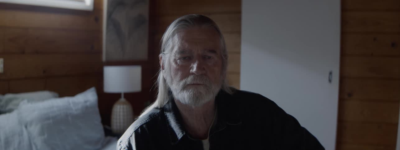 An elderly man with grey facial hair sits on the edge of the bed in his bedroom at midday deep in thought and filled with sadness