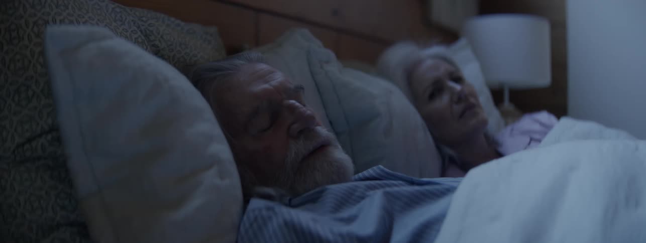 An older man with a white beard and an older woman with white hair are asleep under white covers in a dimly lit bedroom with woodpaneled walls and a lampshade visible on the right