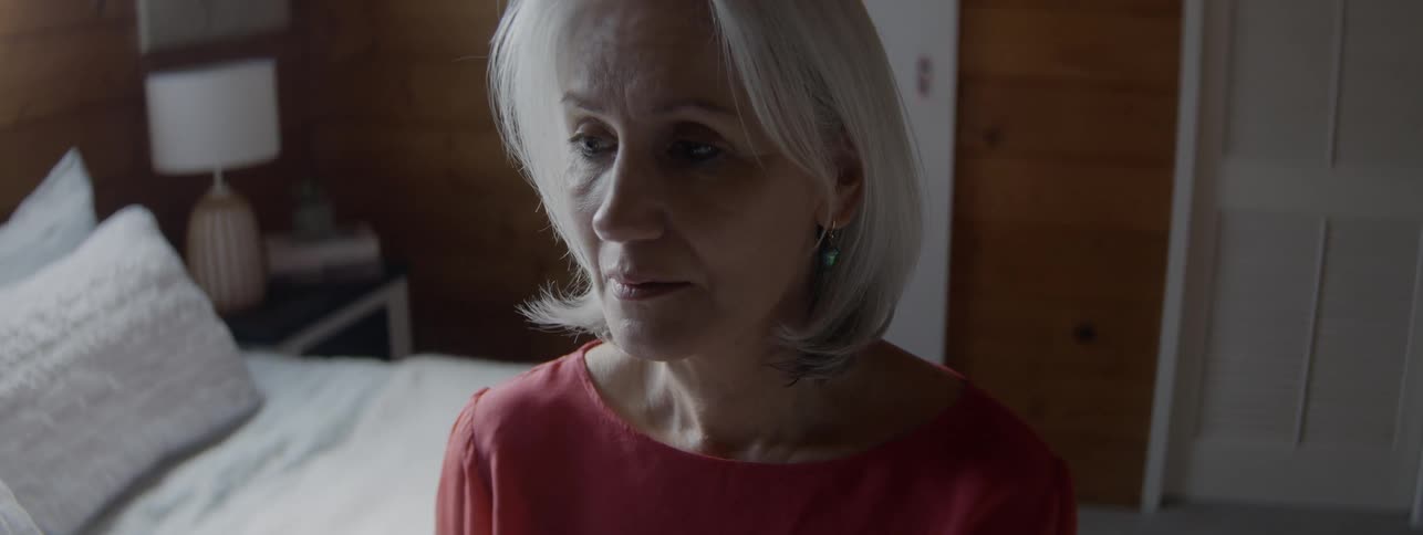 A close view of an elderly woman with short white hair wearing a coral red top shows her looking away with a thoughtful or troubled expression in a dimly lit bedroom