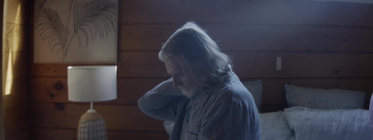 An elderly man with long gray hair and a beard wearing a striped pajama top sits up on a bed and rubs the back of their neck with a woman on the background