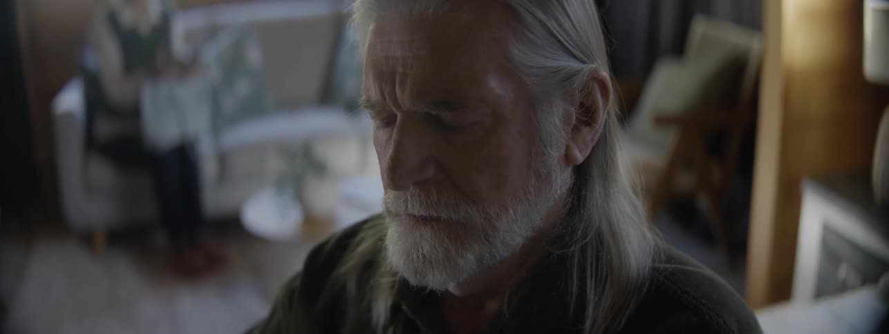 Grayhaired man observes something intently with a focused expression in a cozy room