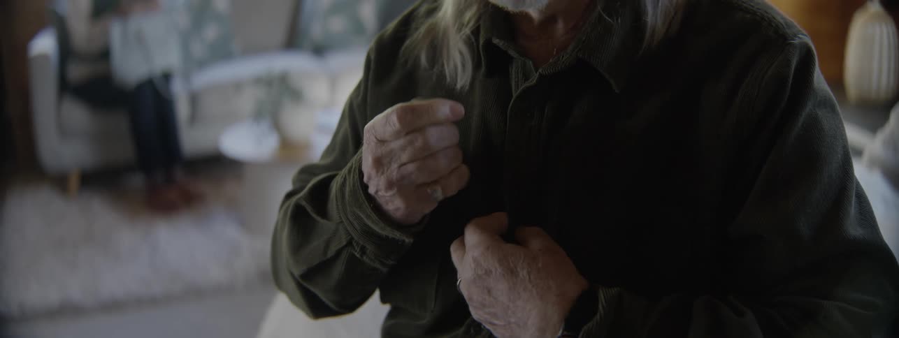 An elderly man with arthritis experiences joint pain while struggling to button up his shirt in the morning