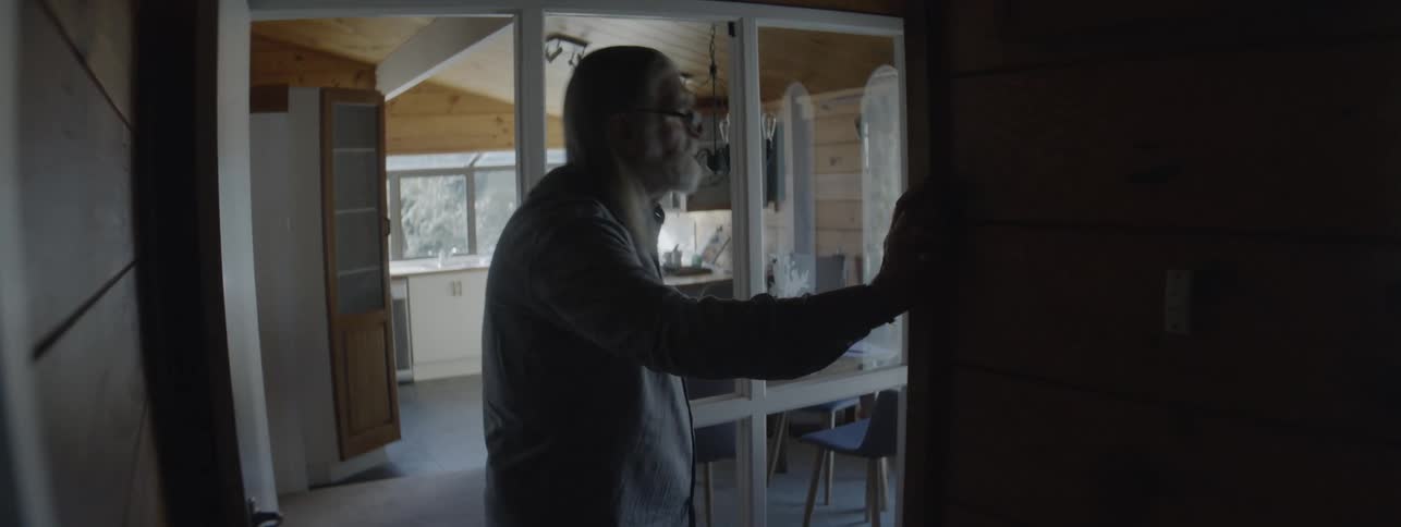 An older man with long hair wearing glasses walks from a brightly lit kitchendining area through an open doorway