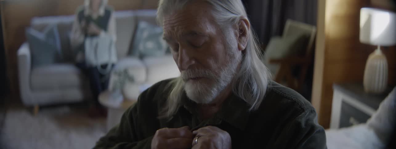 Elderly man buttons up his shirt while getting ready in a room