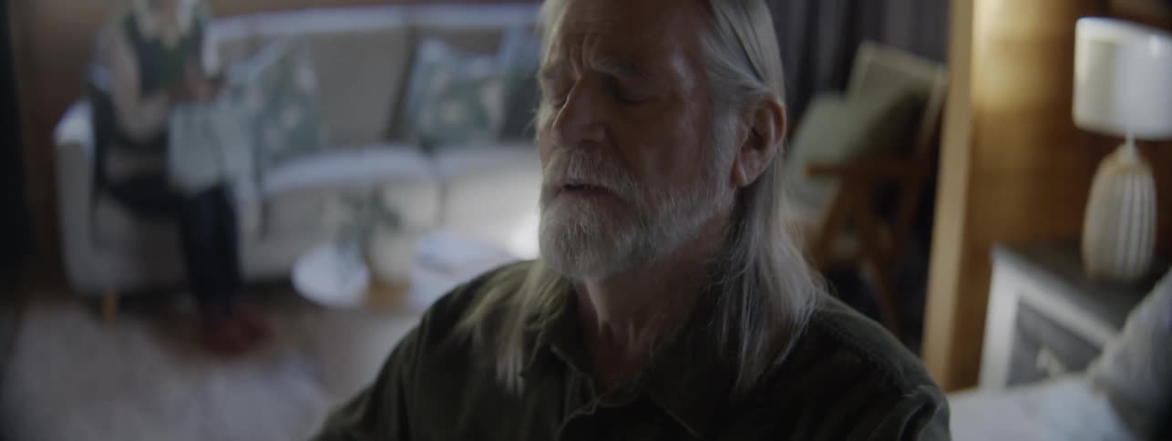 Elderly man buttoning his shirt with a focused expression in a cozy room