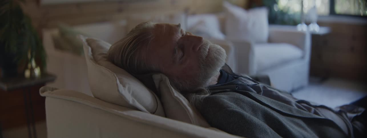 Elderly man rests on the couch with eyes closed in a bright living room