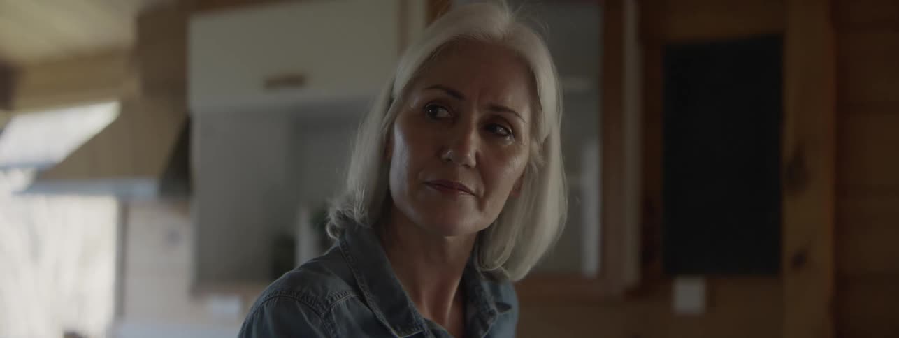 An older woman with white hair wearing a grey shirt picks up a mug and smartphone from a kitchen counter looks at her phone and then smiles thoughtfully in a bright woodpaneled kitchen