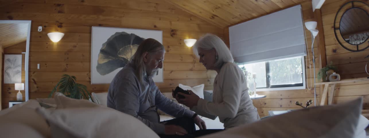 An older man with long hair standing and moving a vacuum cleaner hose who then stops and sits down on a couch at which point an older woman approaches expresses concern touches the mans head and chest and uses a black smartphone to make a call