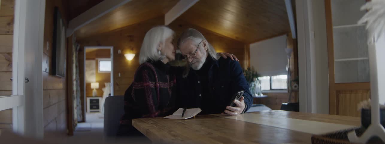 Older couple comforts each other with kisses while viewing a smartphone at home