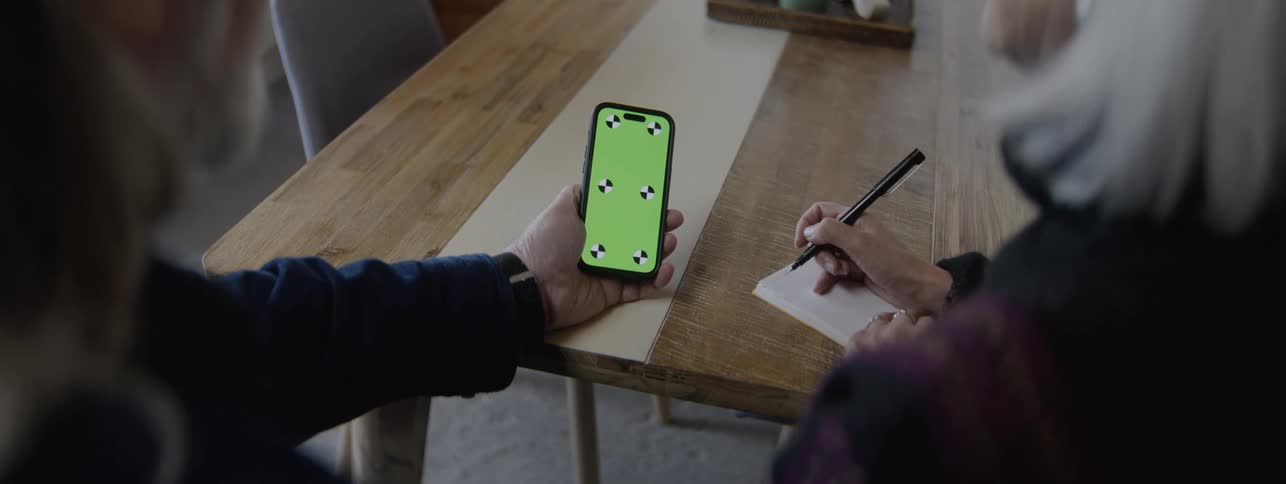 A man holds a smartphone with a green screen and tracking markers as a woman writes on a notepad beside him at a wooden table