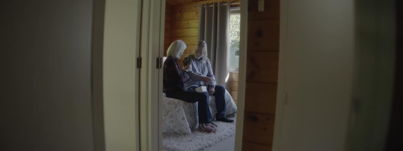 An older woman and older man with grey hair are seated on a bed in a bright woodpaneled bedroom looking down as the woman hands the man a small item or box