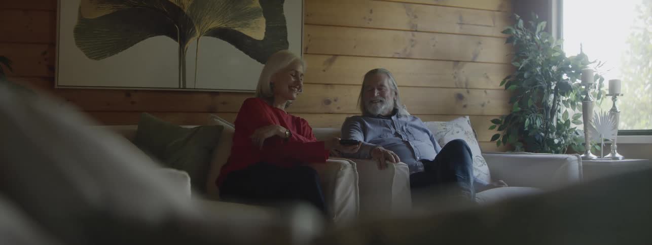 An older man and woman with long gray hair sitting on a couch where the woman interacts with a smartphone and after talking and smiling they both stand up and begin to dance around the living room