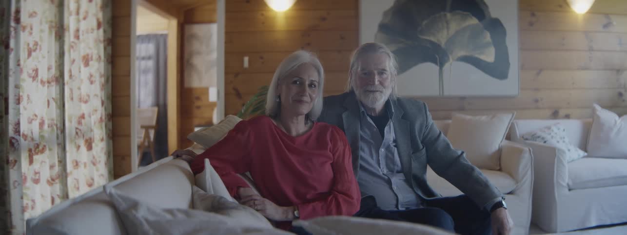 A well dressed older couple smile as they pose together for a photo on a living room couch