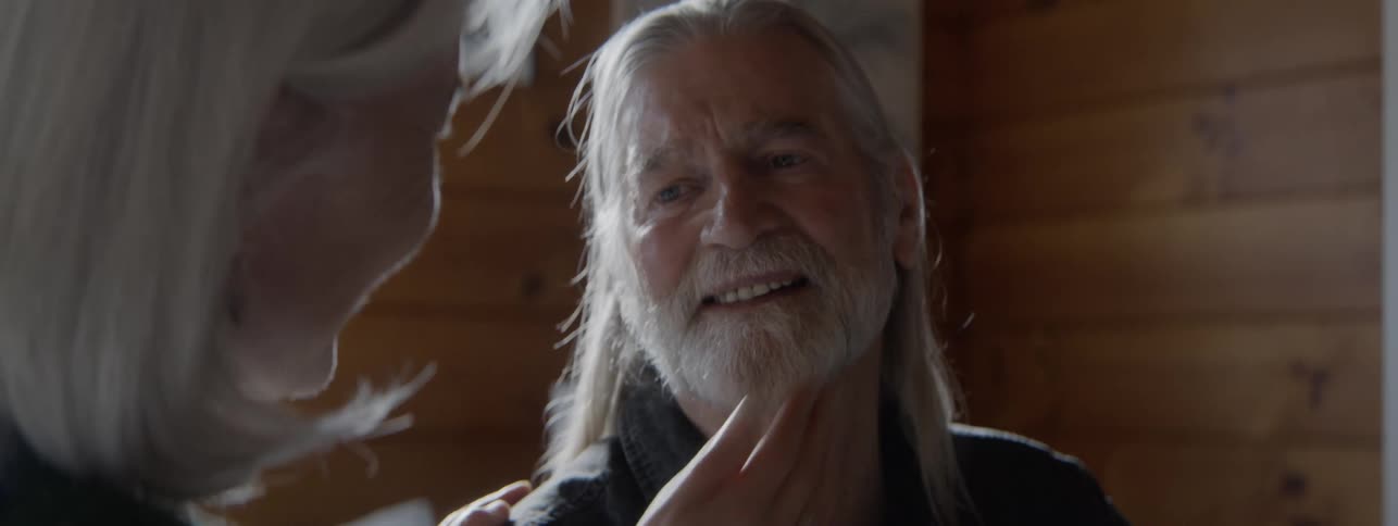 An older man with long gray hair and a beard who is looking at a woman in the foreground while her hand rests on his shoulder and cheek