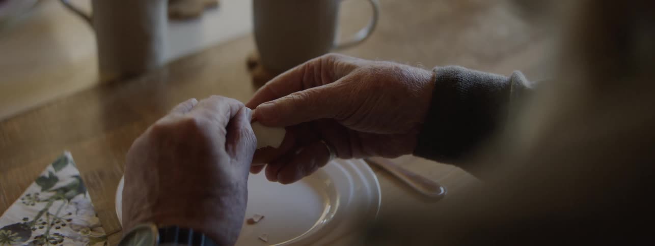 Elderly hands suffering from arthritis struggle to peel a boiled egg over a plate on a breakfast table