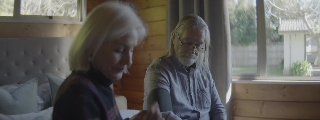 Older woman helps an older man with a blood pressure cuff while sitting in their bedroom at home