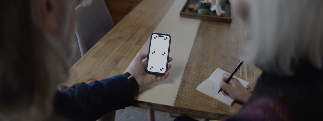 The man holds the cell phone while the woman writes in a notebook