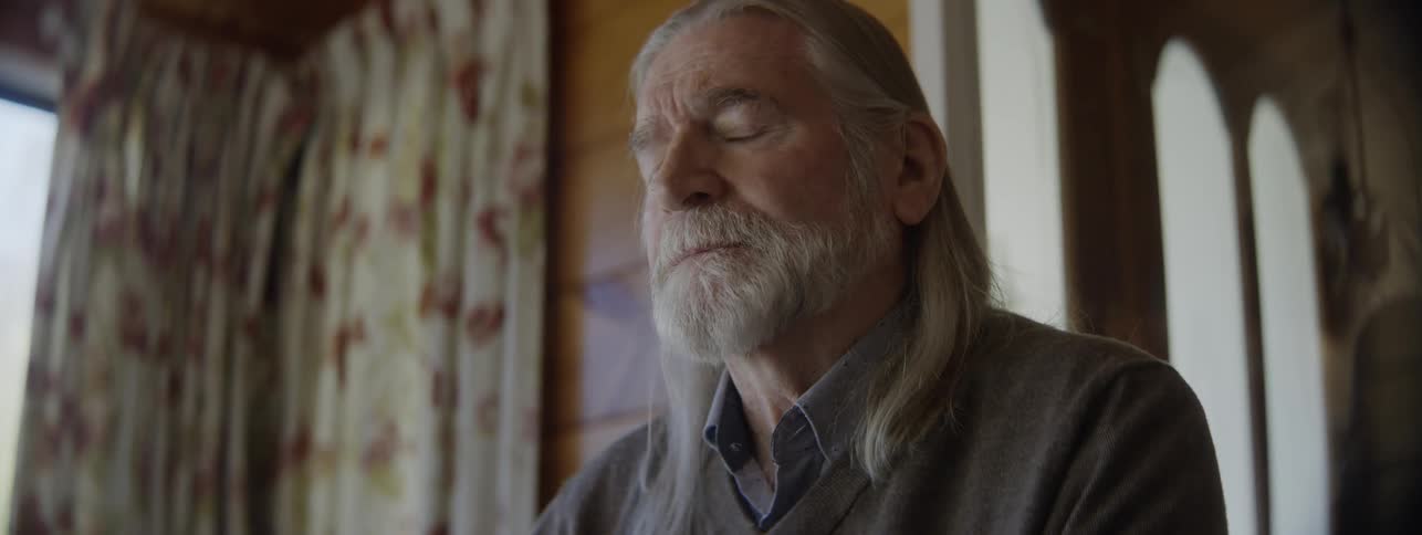 A closeup camera shot focuses on the head and shoulders of an older man with a long gray beard and long gray hair