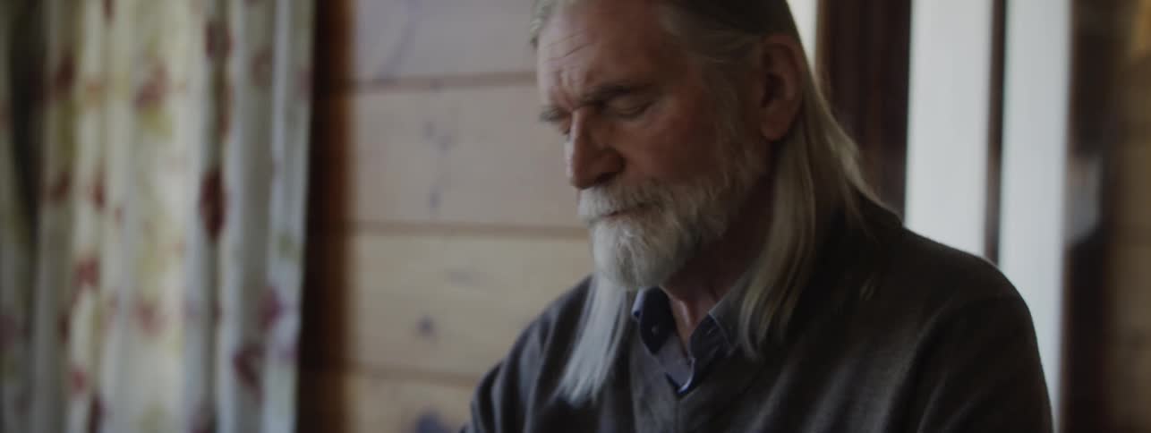 An older man with long grey hair and a white beard wearing a dark sweater peels a hardboiled egg onto a small white plate while seated at a wooden table
