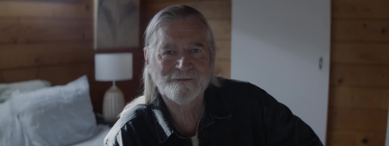 A senior man with long gray hair and a full beard sits in a welllit modern bedroom with wooden walls looking directly into the camera before a slight smile appears on his face