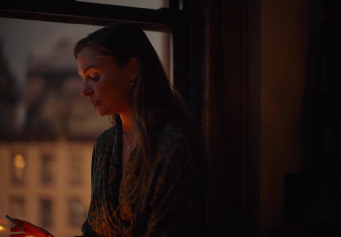 A person holding a glass jar with a lit wick inside and the person then turns to show the face of a woman with long hair who raises her head and looks out a window with buildings in the background