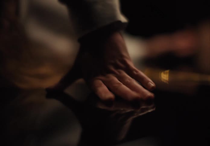 A close dark view shows the hand of a woman in a light sleeve reaching out to touch a dark reflective surface as she leans forward and her hair falls around her face