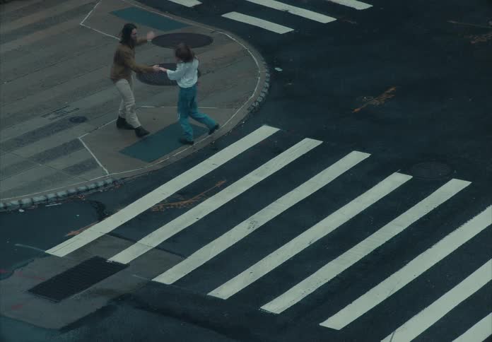 An overhead camera view shows a man and a woman standing on the curb of a street corner near a crosswalk where they hold hands raise their arms and dance playfully while a man cross the street and a dark vehicle drives through the lower foreground