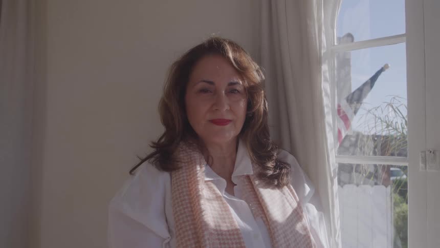 A woman with long brown hair wearing a scarf and a white shirt standing and looking directly ahead framed by a window with a flag visible outside