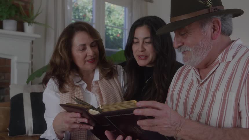 An older man wearing a hat an older woman and a younger woman all gathered around a book in a living room setting